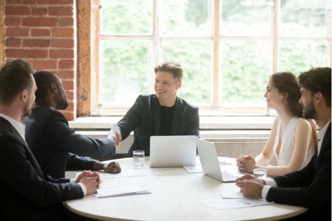 A committee gathered at a table, with one member shaking hands with a successful recruit as the others smile on.